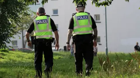 Alan Simpson Two police officers with their back to the camera. Both are wearing dark clothing and boots with hi-viz jackets over the top with the word police on the back in white writing on a blue background. The officer on the right is wearing a cap. They are walking through long green grass towards a white block of flats with small windows in the side.