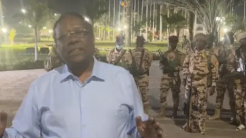 Facebook Foreign Minister Abderaman Koulamallah seen in a video apparently shot at the presidential palace in N'Djamena. He is wearing a blue shirt. Behind him are lines of palm trees and a lawn. Over his shoulder is also a group of soldiers in camo uniforms. 