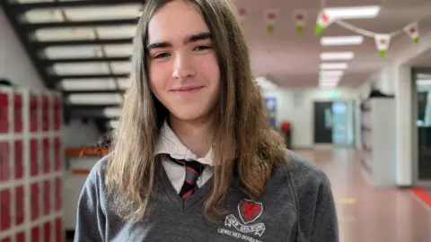 Alexia smiles into the camera. She is wearing a grey school jumper and has long brown hair. She is standing in a school hallway, with red lockers, stairs and Welsh flag bunting in the background. 