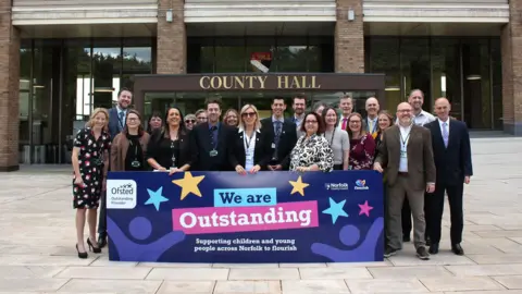 Norfolk County Council A group of people stand in front of Norfolk County Council's County Hall building in Norwich, holding a blue-coloured sign saying 'We are Outstanding'.