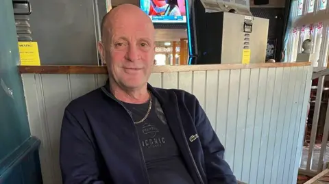 An archive photo of John Smith who is sitting at what appears to be a pub. He is wearing dark clothes and a gold chain around his neck. He is looking at the camera and smiling.