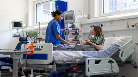 A female nurse in blue scrubs speaks to a female patient lying in a hospital bed.