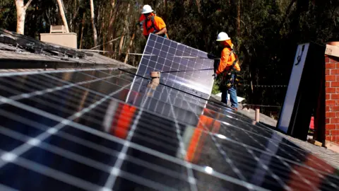 Two workers wearing high viz jackets and hard hats installing solar panels on a building's roof.