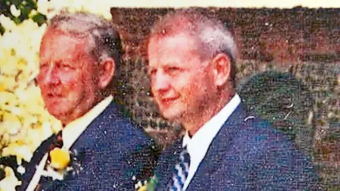 PA Media An old photo of Conrad Wright and his son Steve Wright at a family wedding. They both are looking away from the camera and are wearing navy blue suits with white shirts and ties. 