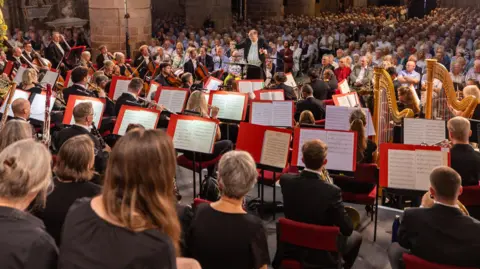 Dale Hodgetts An orchestra performing in front of a seated crowd inside Gloucester Cathedral. It shows a number of musicians sat in front of music stands playing instruments, including two harms, clarinets, violins and other string and wind instruments.