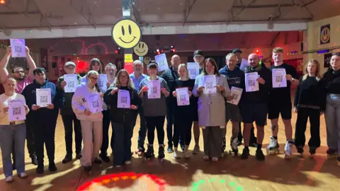 A large group of people in the roller rink holding pieces of paper reading: 'Help to save Wigan Roller Rink'.