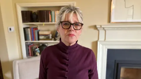 Mark Ansell/BBC Pauline McLeod, who has blonde hair and black-rimmed glasses. She stares directly at the camera with a neutral expression.