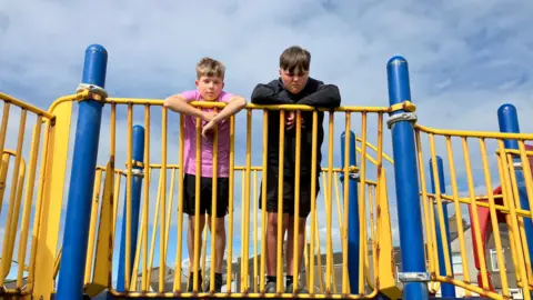 Ethan and Kaelan standing on a piece of play equipment. They are leaning on the yellow railing and looking down at the camera. They look unhappy. Red and brown rust can be seen in many places on the equipment.