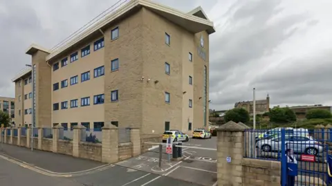 Google A four-storey modern beige brick building. Police cars are parked in the car park outside it, which is barriered off.