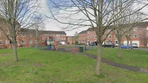 Google Maps A children's playground pictured in the middle of a housing estate. The playground is surrounded by grass with several trees. There are three storey red brick buildings around the edge of the grass.