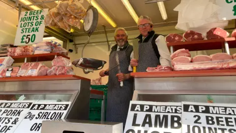 Two men wearing aprons, standing inside a van which is selling meat. Shelves in front of them are full of joints of meat.