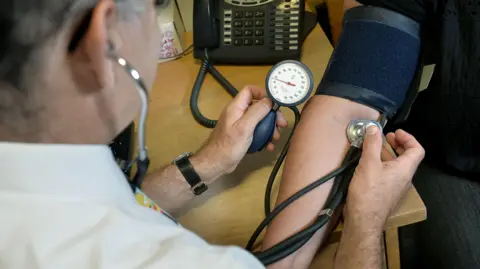 A male doctor wearing a white shirt is photographed from behind while he uses equipment to check a patient's blood pressure.