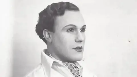Maurice Dobson Museum A black and white photo of a young man in profile looking to the right. He has slicked back dark curly hair and is wearing lipstick and eye make up. He's dressed in a white shirt and is wearing a cravat with a geometric pattern