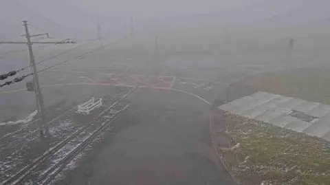 The remains of some snow on the ground at the tram crossing at the Bungalow on the A18. It is foggy and visibility is poor.