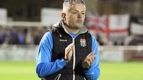 Bath Chronicle Jerry Gill on the pitch wearing a bright blue sweat shirt and black Bath City gillet, looking solemn and clapping his hands