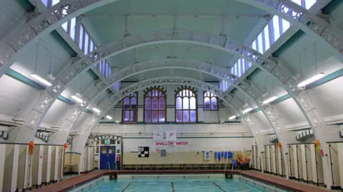 Getty Images The pool in 2005 filled with water. There are cubicles on either side of the pool and white metal arches on the roof. There are windows on the back wall and there are blue double doors and a sign that reads "shallow water"