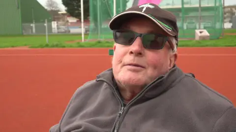 An older man with a brown baseball cap and a grey fleece, wearing dark glasses.