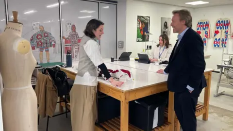 Two students talk to the Vice Chancellor Professor Paul Fieldsend-Danks of the Arts University Plymouth. He is dressed in a dark blue suit. They are leaning over a table containing a white jacket that they are making for their degree course. There is a tailor's dummy in the foreground and clothing from a previous project hanging on the walls behind in the Fashion Studio.