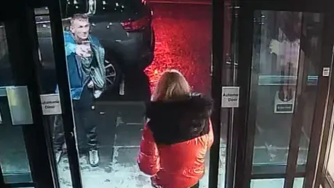 COPFS CCTV image of of Michael Harvey shouting at Carol O'Reilly at sliding doors of a a hotel. She is wearing a black and red jacket and he is wearing a blue jacket and is pointing at her