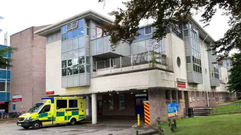 An ambulance waits outside a covered back entrance at an aging hospital, which has marks on its white render. It is about three stories tall with windows jutting out, and there is a green grass verge to the right.