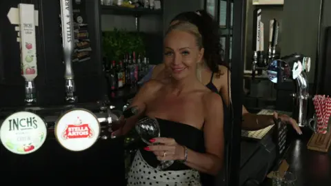 A woman standing behind a bar in a pub, holding a stemmed glass in one hand and a wine bottle in the other. In the foreground, beer taps display labels including Inch’s Cider and Stella Artois. Behind the counter are shelves stocked with bottles of wine and spirits, along with a green plant wall feature. Another person is partially visible behind her. On the right, a countertop holds a container of red-and-white striped straws.