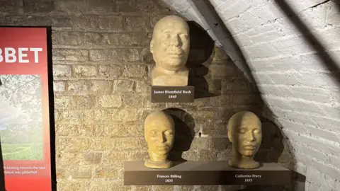 Emma Craig/BBC Three 19th Century death masks on plinths against a brick wall in a basement. The death masks are yellow in colour and are labelled James Blomfield Rush, Frances Billing and Catherine Frary.