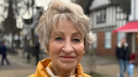 An elderly woman with short grey hair and wearing a yellow coat standing in the middle of a high street.