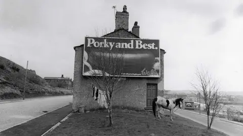 Ian Beesley A black and white photo of a horse tethered on grass in front of the gable end of a house with an advertising poster on it with the slogan 'Porky and Best'. 