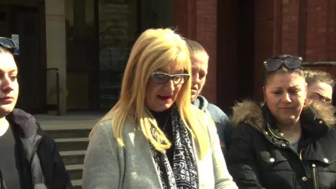 Karen Dry (centre) has long blonde hair and glasses. She stands outside a brick building and her head is bowed down as she reads from a piece of paper, while several people stand closely around her. She wears a light grey coat over a black top and patterned scarf. The group is gathered on the steps of Hull Crown Court in bright sunlight.