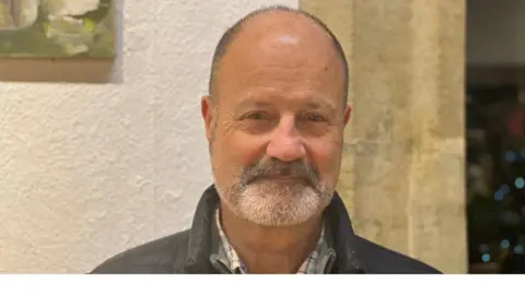 Cranleigh Arts Brian Freeston, a bald man with a grey beard and wearing a checked shirt and brown jumper, looks straight into the camera.