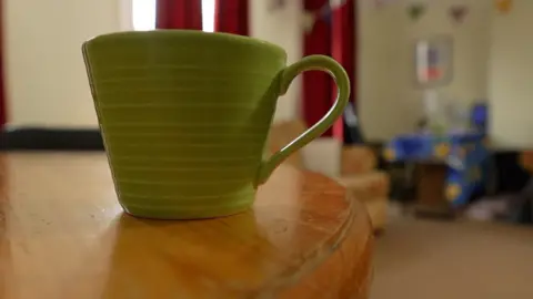 Steve Hubbard/BBC A green mug sitting on a wooden table. Windows, red curtains and a sofa can just be made out in the background.