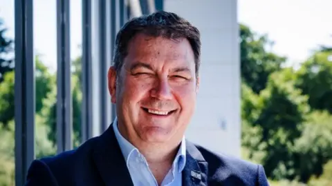 BBC Steve Turner. He has brown hair and is smiling widely. he is wearing a blue suit with a white shirt. There is a union flag pin on his lapel.