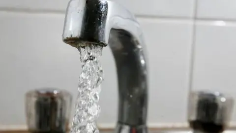 A silver tap pouring out water. White tiles are in the background.