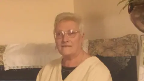 Family handout Noreen is sat inside a house on a patterned sofa. She has short grey hair and glasses. She is wearing a white top.