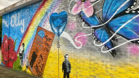 A wall mural that includes a young schoolboy, a blue balloon shaped like a heart, a blue butterfly, a rainbow, and a red rose