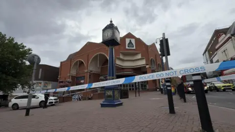 Blue and white police tape surrounds a tall blue clock outside a brick-built shopping centre. There are police vehicles parked either side of the cordon and a fire engine visible too. The scene is ser against a grey cloudy sky.