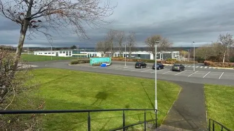 BBC Prudhoe High School viewed from the top of steps into the school groups is a one storey building in white and grey cladding set over a large area. In front is a carpark around the school are playing fields 