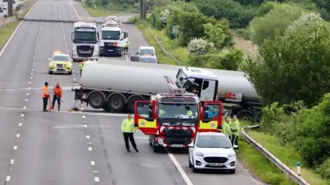 BBC An empty motorway with a truck across the road and into the barrier and hedge. There is a fire truck and emergency services in high vis jackets.