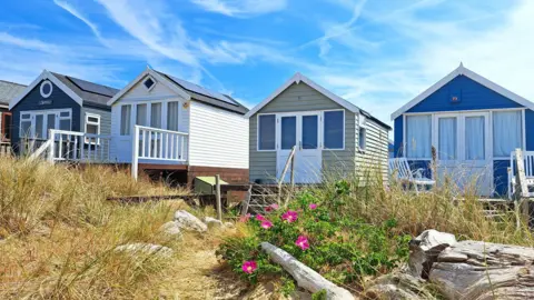 MarkieB A row of four beach huts (dark blue, white, grey/green, royal blue) with a blue sky criss-crossed by wispy cloud behind, and beach-type scrub in front