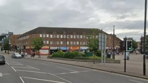 Drummond Street - a busy street with multiple lanes of traffic and several buildings lining the roadside. A few cars are driving along the road, and some pedestrians can be seen walking on the pavements. In the centre of the image, a large brick building with shops on the ground floor sits at the corner of the junction, with trees and a small landscaped area in front of it.