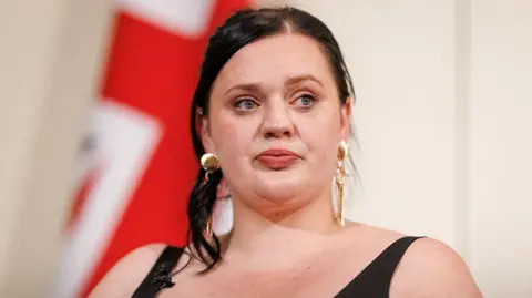 PA Media A woman with dark brown hair wearing a black vest top. Her hair is tied in a ponytail and she is looking to the side. She is wearing gold earrings. There is a Union flag behind her.
