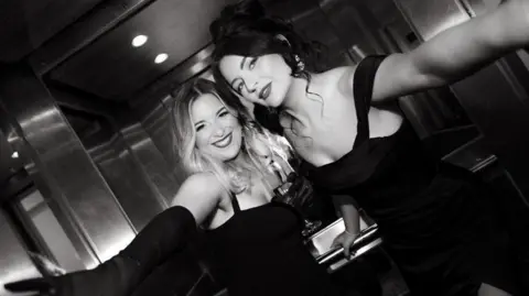 The Unfiltered Bride Georgie Mitchell and Beth Smith are pictured here in a lift. The photograph is black and white. Georgie has long blonde hair and wears a black dress. Beth has dark hair which is pinned in a bun. She too is wearing a black dress. 