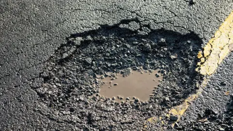 A close up of a pothole on a double yellow lined road with muddy water in the bottom