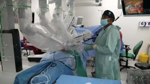 A robot with several arms performing a surgery in an operating theatre. There is a surgeon in a green frock coat who is wearing a face mask. There is a monitor in the background and surgical equipment in the foreground.