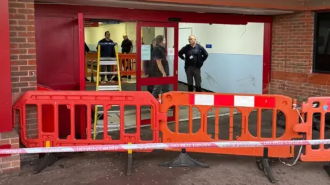 Eddie Mitchell Orange barriers block off a large door to the building. Staff can be seen inside 