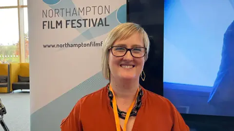 Ollie Conopo/BBC Becky Carrier with medium-length blond hair wearing an orange top and lanyard. A sign behind her says "Northampton Film Festival".