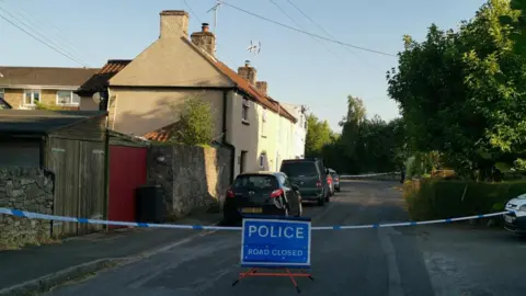 A quiet residential street, with a few houses along one side and trees on the other, is blocked by police tape and a sign which says 'Police, road closed'.