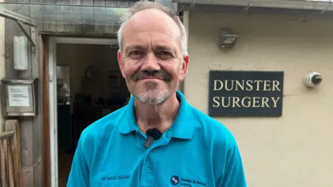 A gp called Dr David Davies stands outside his surgery in Dunster. He has a  short beard and moustache and is wearing a turquoise polo top