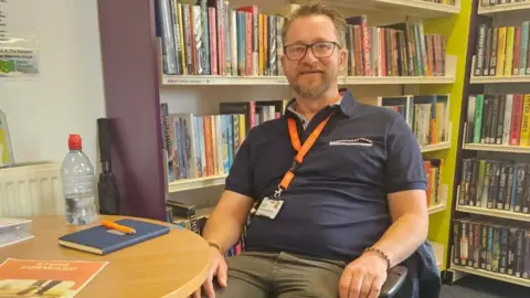 Jimi Wilson wearing a navy polo shirt sits in a chair in a library. He has an orange lanyard around his neck and is setting in front of two bookcases. On the table in front of him is a notepad, a bottle of water with a red cap, a white binder and an orange leaflet.
