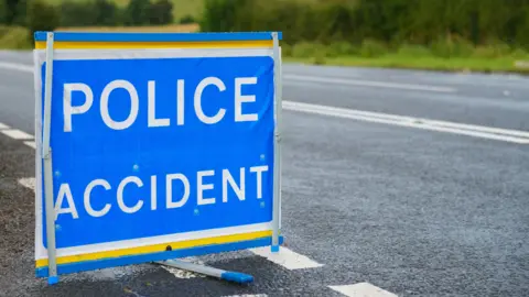 A blue sign at the side of the road which says 'POLICE ACCIDENT' on it. An empty road can be seen in the background, with a hedgerow in the distance. 